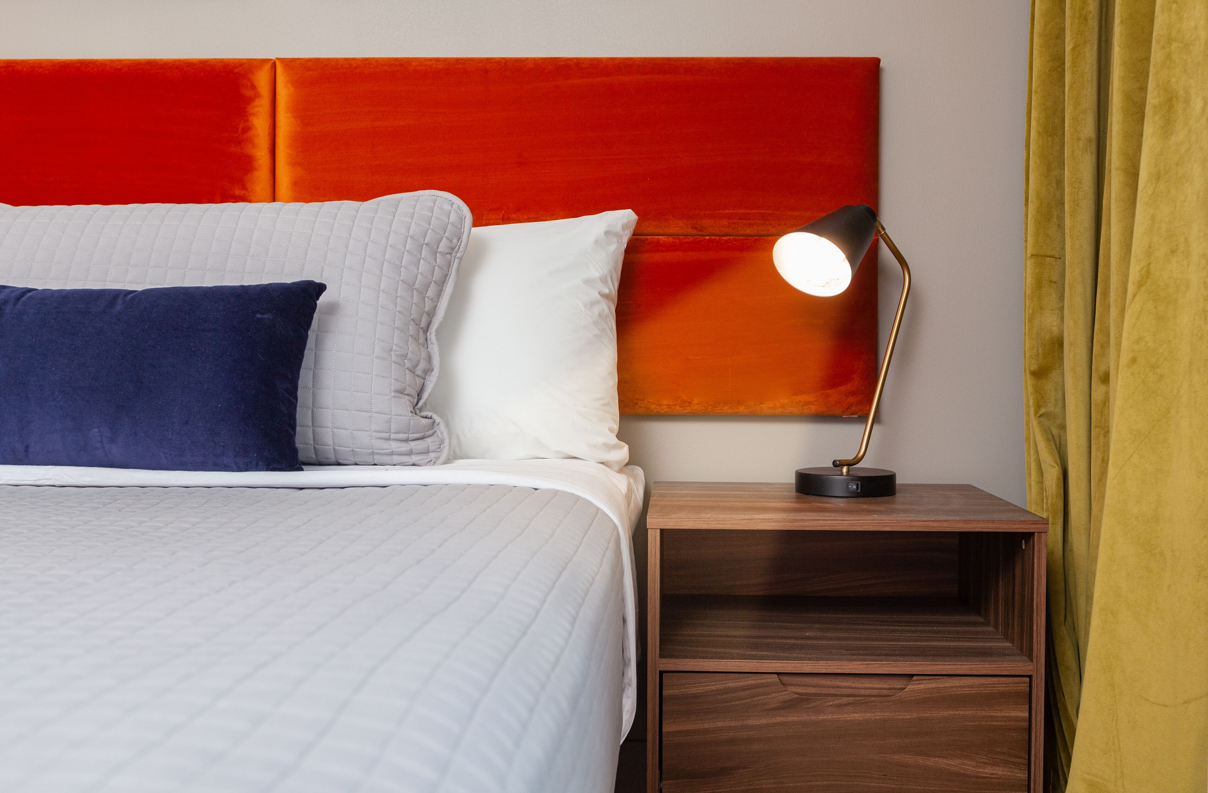 Een net opgemaakt bed met een wit en blauw kussen, een rode hoofdbord en een nachtkastje met een lamp.
