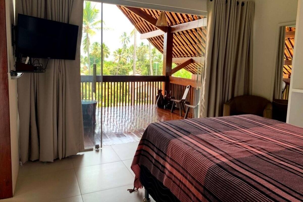 Une chambre d’hôtel avec un lit, une télévision, un balcon agrémenté d’une pergola en bois et une vue sur des palmiers.