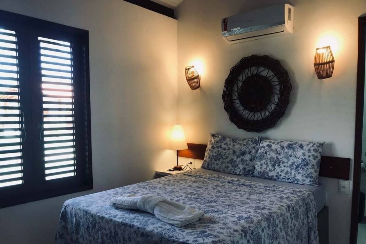 Une chambre à coucher comprenant un lit, des lampes de chevet, un climatiseur fixé au mur et une fenêtre avec des stores.