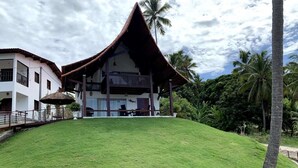 Un edificio con techo inclinado y balcón, rodeado de abundante follaje y palmeras.