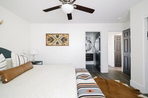 A bedroom with a bed, a ceiling fan, a wall-mounted artwork, a nightstand, and a door leading to a bathroom.