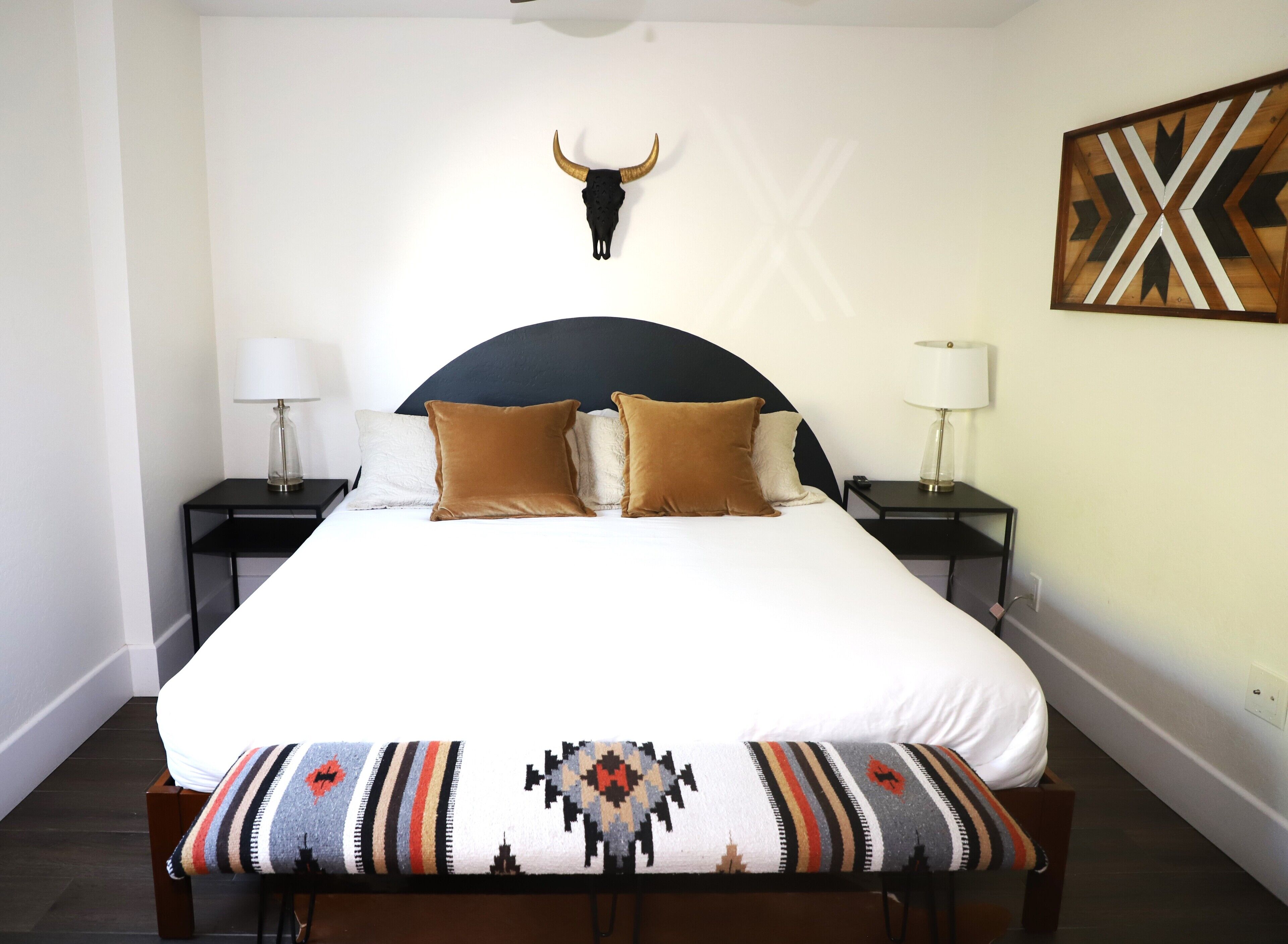 Un letto ben rifatto con un piumone bianco, due cuscini marroni e una coperta decorata. Si nota una testiera nera, due comodini con lampade e una decorazione a teschio di toro fissata al muro.