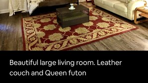 A large living room with a leather couch and a Queen futon, featuring a red and gold patterned rug, wooden flooring, and a ceiling fan. - BestStays@Flowers Near UT Tyler large groups welcome (Tyler)