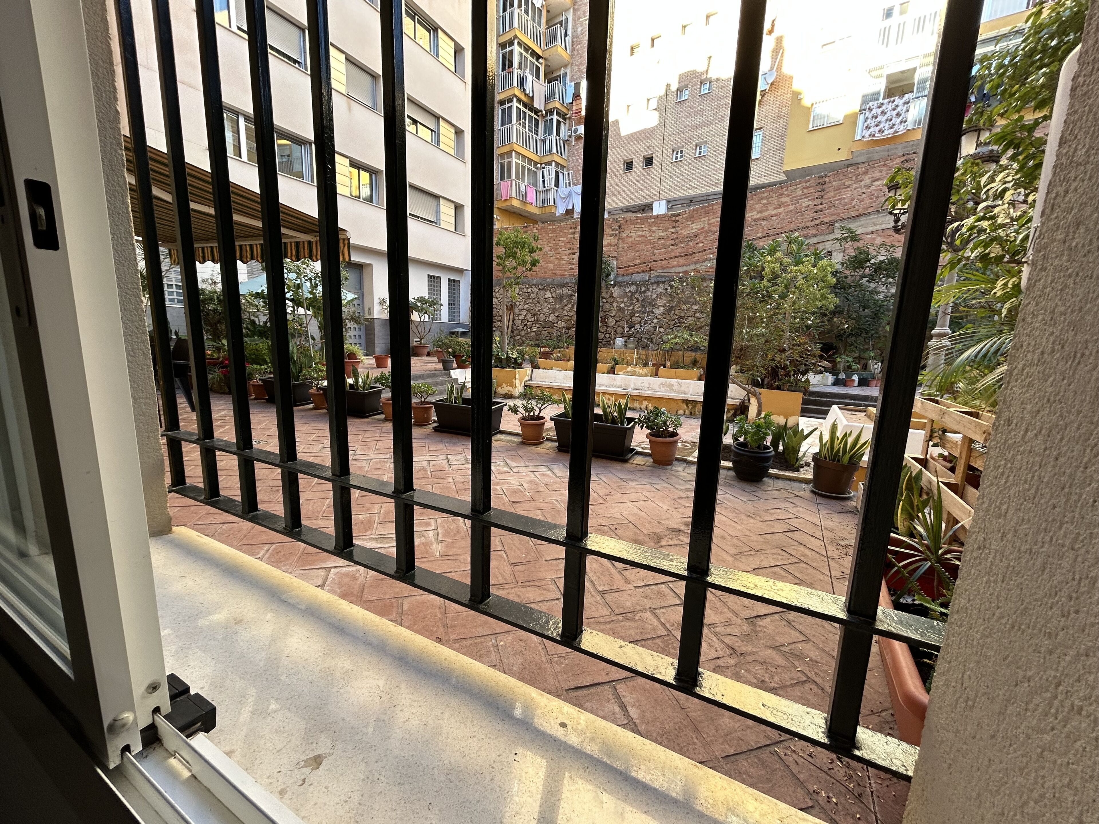 A courtyard with potted plants, a stone wall, and a building with balconies in the background.