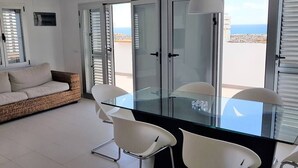 A modern dining area with a glass table and white chairs, a wicker sofa, and a view of the sea through open doors. - Casa La Vista - Bungalow with fantastic sea views (Mogan)