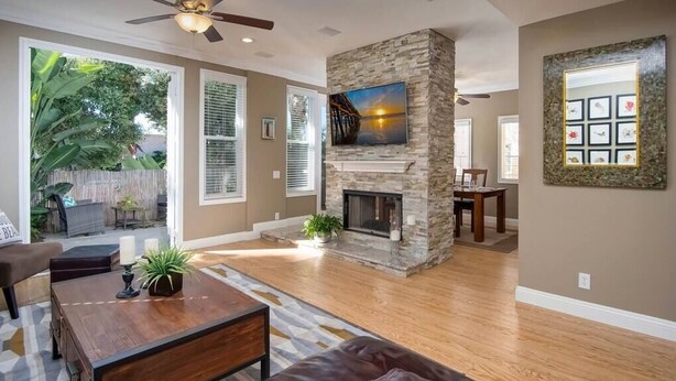 A spacious living room with hardwood flooring, a stone fireplace, a ceiling fan, and a large window overlooking a backyard.