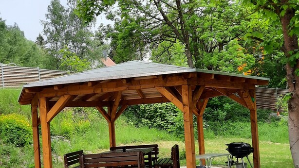 A wooden gazebo with benches and a barbecue grill in a grassy area.