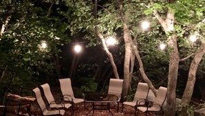 An outdoor seating area with chairs and a table, surrounded by trees and illuminated by string lights.