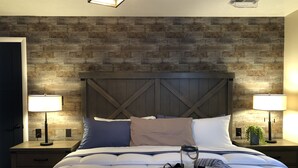 A neatly made bed with pillows, a bedside table with a lamp, a nightstand with a plant, and a rustic wooden wall.