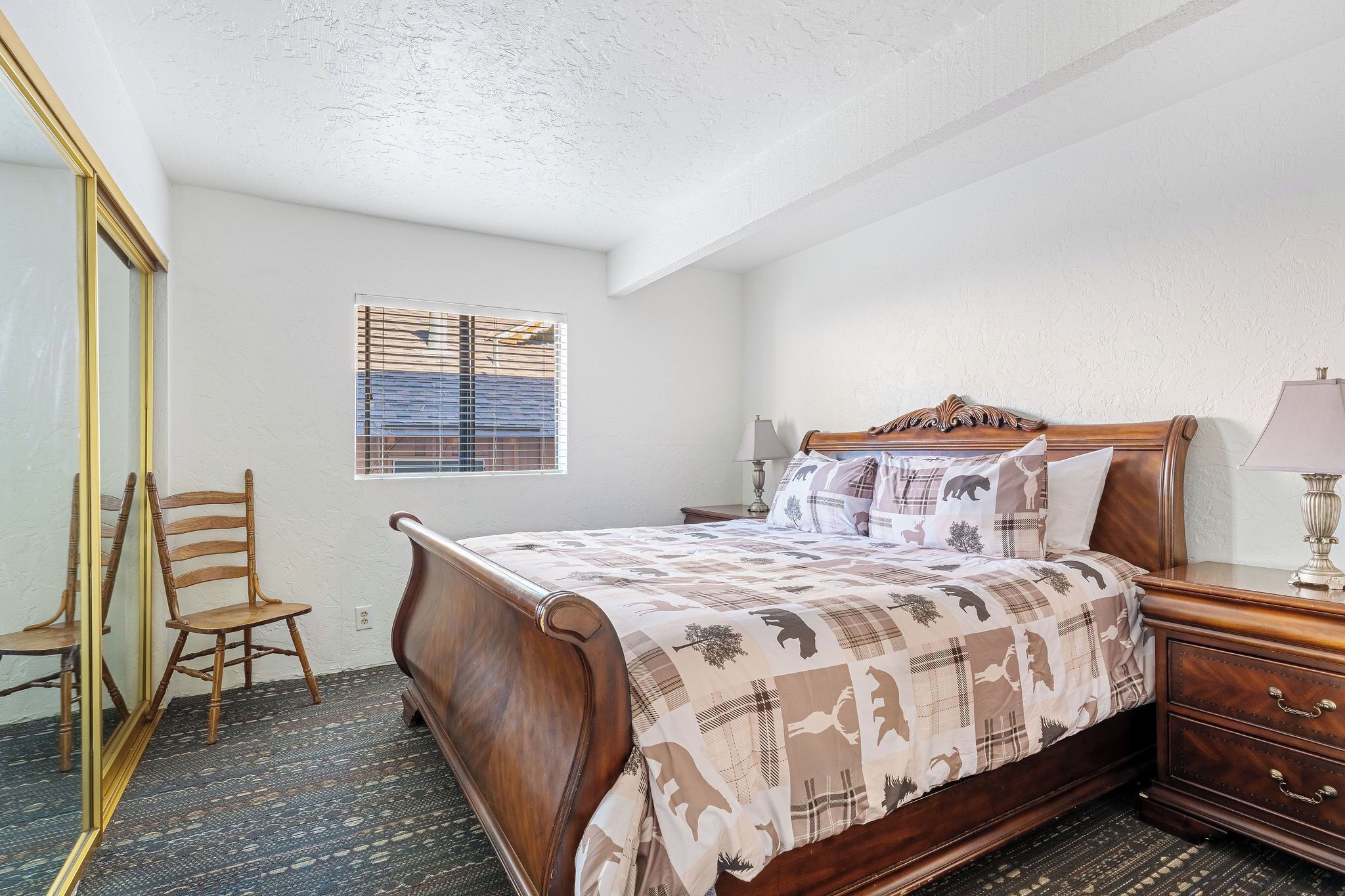 A bedroom with a wooden bed, a nightstand, a mirror, a window with blinds, and a chair.
