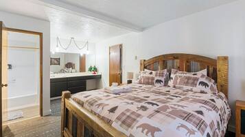 A bedroom with a wooden headboard, a bed with a bear-themed duvet, a bedside table, a nightstand, a wooden door, a bathroom with a mirror, and a small plant.