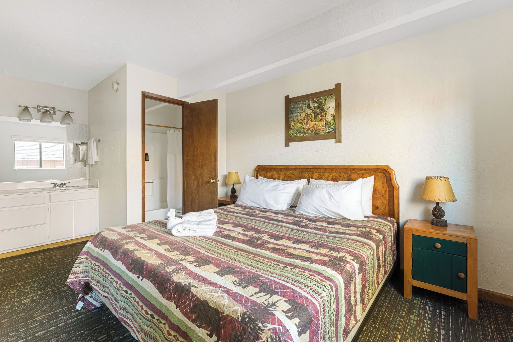 Een hotelkamer met een groot bed, een houten nachtkastje met een lamp, een klein kastje, een badkamer met een wastafel en spiegel, en een schilderij aan de muur.