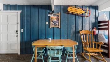 A dining area with a wooden table and chairs, a wooden rocking chair, a framed picture on the wall, and a ski lodge sign.