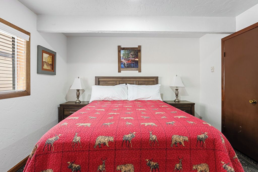 Quarto com edredom acolchoado vermelho com estampas de animais, duas luminárias de cabeceira, cabeceira de madeira, um quadro na parede e uma porta à direita.