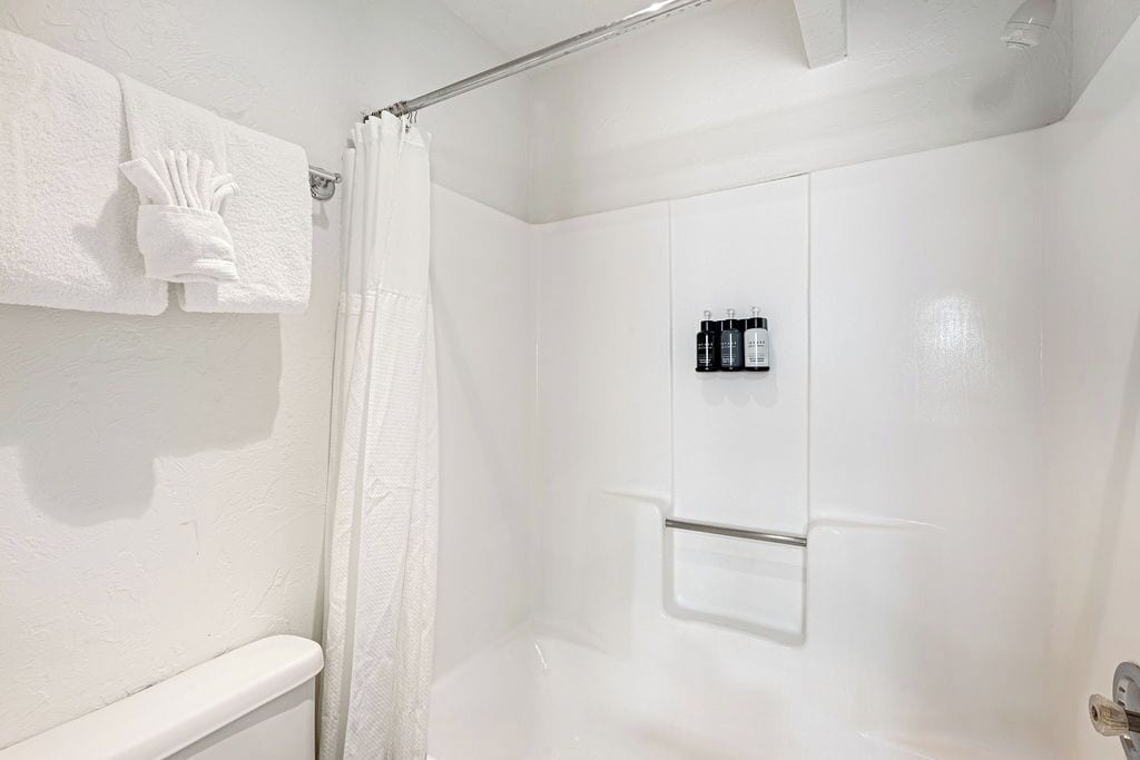 A white bathroom with a shower curtain, a towel rack with towels, and a shelf with toiletries.