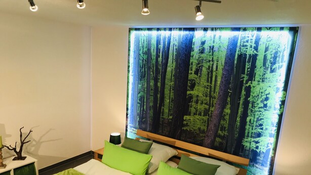 Een moderne slaapkamer met een groot bed, groen beddengoed en een behang met een bosmotief.