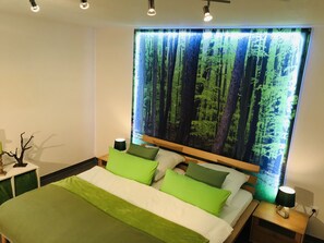 Un dormitori modern amb un llit gran, roba de llit verda i un mural de paret amb motius de bosc.