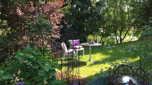Een tuin met een ronde tafel en twee stoelen, omringd door diverse planten en een decoratieve metalen bol.