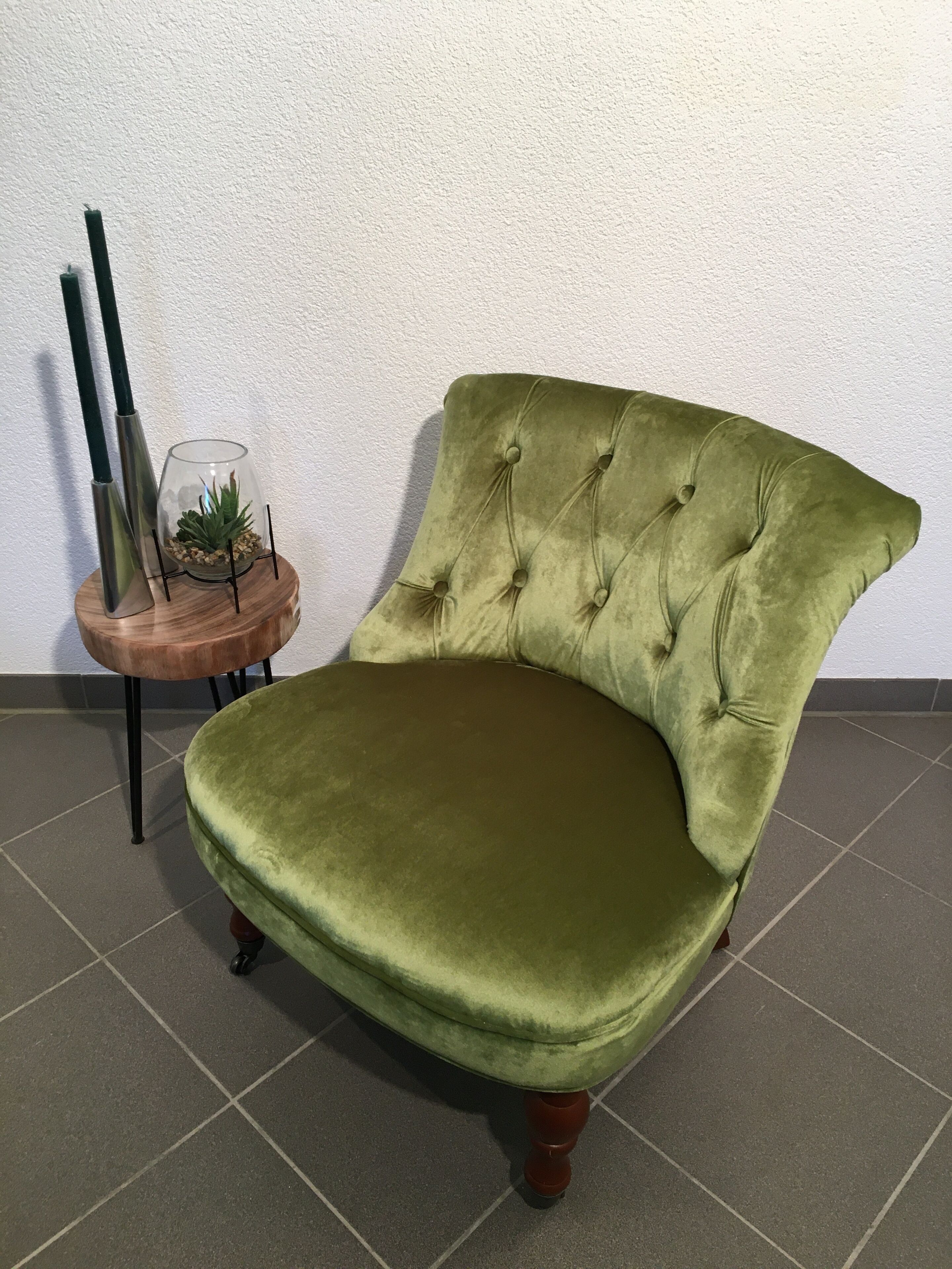 En grøn velour-lænestol med en knapryg, placeret på et flisegulv ved siden af et lille træ-sidetæppe, der holder en glasvas med en plante.
