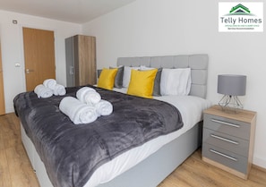 Une chambre à coucher moderne avec un lit gris, des oreillers blancs et jaunes, une tête de lit grise, une armoire en bois et une table de chevet avec une lampe.
