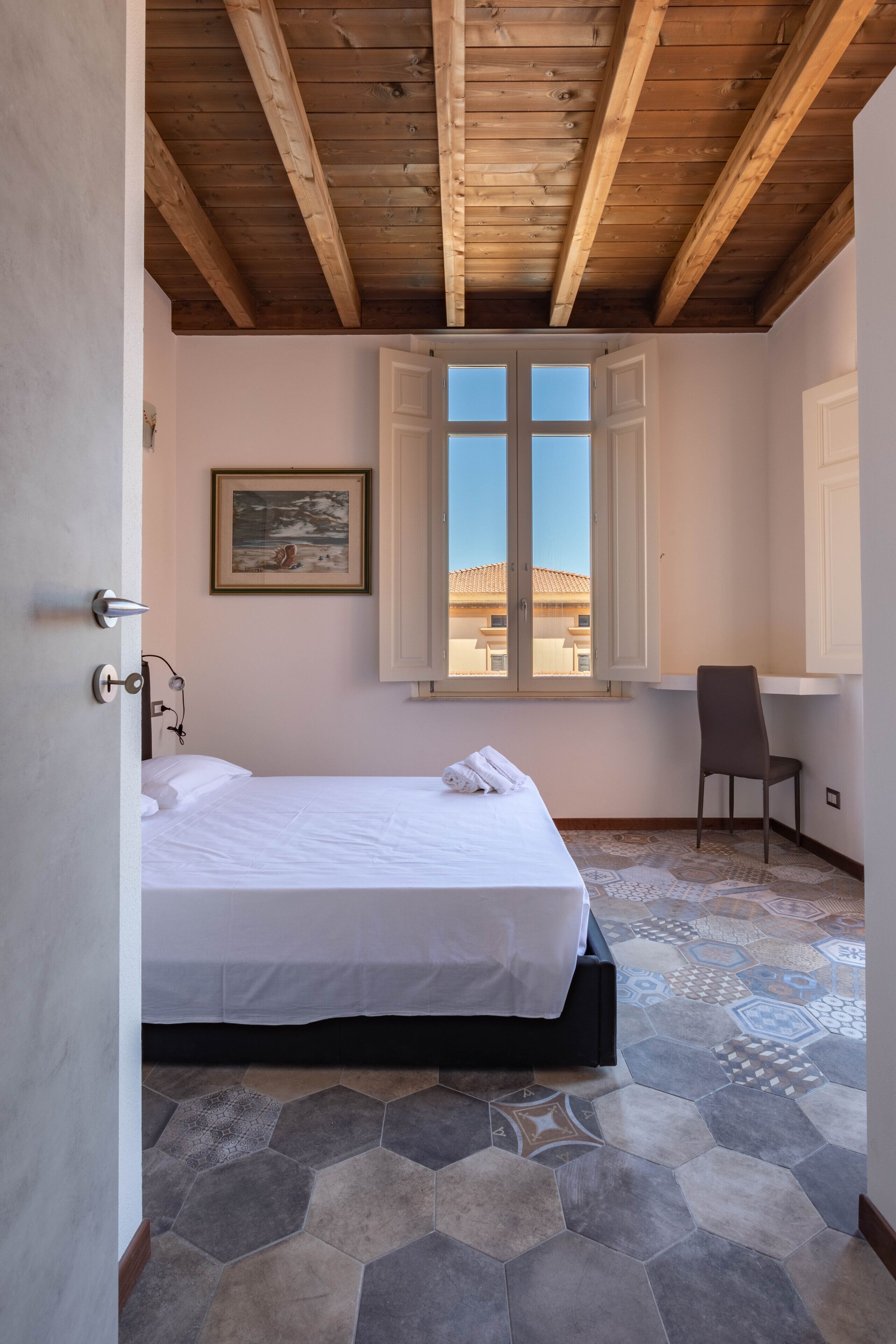 Un dormitorio con una cama, una silla, una ventana con contraventanas abiertas y un cuadro en la pared.