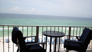 A balcony with two chairs and a small table, overlooking a beach with umbrellas and people.