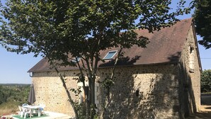 Ein Steinhaus mit einem Ziegeldach, einer kleinen Terrasse mit Gartenmöbeln und einem Baum davor.