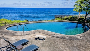 A swimming pool with a curved edge, surrounded by stone tiles. There are two lounge chairs and a small table. The pool is adjacent to a rocky shoreline with a view of the ocean. - Hilo Shangrila - Oceanfront, Pool and Hot Tub Private Vacation Rental (Keaau)