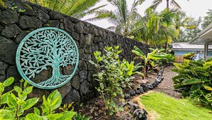 A stone wall with a decorative tree metalwork, lush tropical plants, and a pathway leading to a building. - Hilo Shangrila - Oceanfront, Pool and Hot Tub Private Vacation Rental (Keaau)