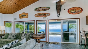 A spacious living area with a large sofa, a coffee table, and a view of the ocean through sliding glass doors. There are surfboard-shaped wall decorations and a ceiling fan. - Hilo Shangrila - Oceanfront, Pool and Hot Tub Private Vacation Rental (Keaau)