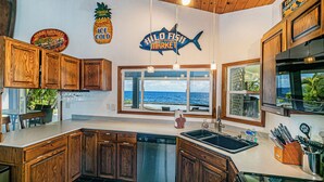 A kitchen with wooden cabinets, a dishwasher, a large window with a view of the ocean, and a fish-shaped sign. - Hilo Shangrila - Oceanfront, Pool and Hot Tub Private Vacation Rental (Keaau)