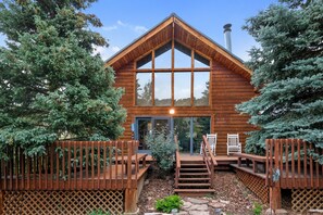 Situated in a wooded area, this cabin features a gabled roof and large windows, with a wooden deck and stone pathway leading to the entrance.