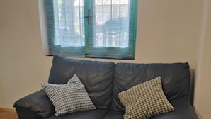 A dark leather sofa with two patterned cushions, a window with green blinds, and wooden flooring. - Two Bedroom Apartment (Bordighera)