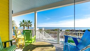 A covered outdoor area with a wooden deck, green and blue Adirondack chairs, a round wooden table, and a view of the ocean. - Casa Banana Oceanfront on Ocean Blvd, 6 BD, sleeps 20, pool,hot tub, elevator (Isle Of Palms)