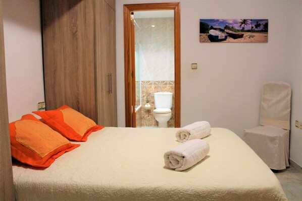 A hotel room with a bed, orange pillows, a white bathroom with a toilet, and a small white chair. - APARTMENT 2 MINUTES FROM PUERTA DEL MAR BEACH (Almuñécar)