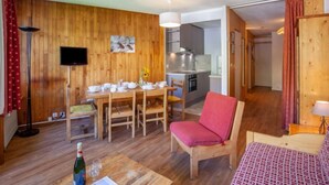 A wooden-floored living area with a dining table set for four, a red chair, and a sofa. A kitchen is visible in the background. - Le Curling B (Val Claret) by Interhome (Tignes)