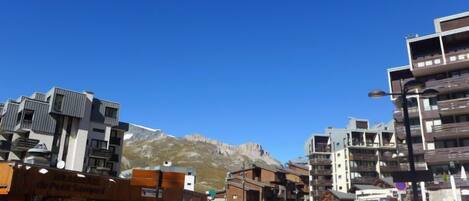 Une rue moderne en milieu urbain, bordée d’immeubles à plusieurs étages, avec un ciel bleu dégagé et une montagne en arrière-plan.
