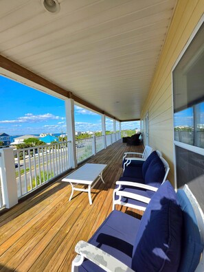 Une terrasse extérieure couverte avec vue sur l’océan, dotée d’un plancher en bois, de rampes blanches et d’un espace salon avec des coussins bleus.