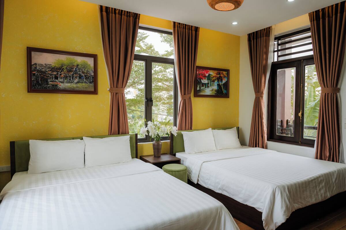 A hotel room with two beds, a small table with a vase of flowers, and two paintings on the walls.