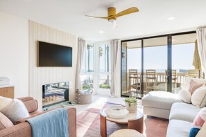 A spacious living room with a flat-screen TV, comfortable seating, a ceiling fan, and a large sliding glass door leading to a balcony with a view of the ocean.