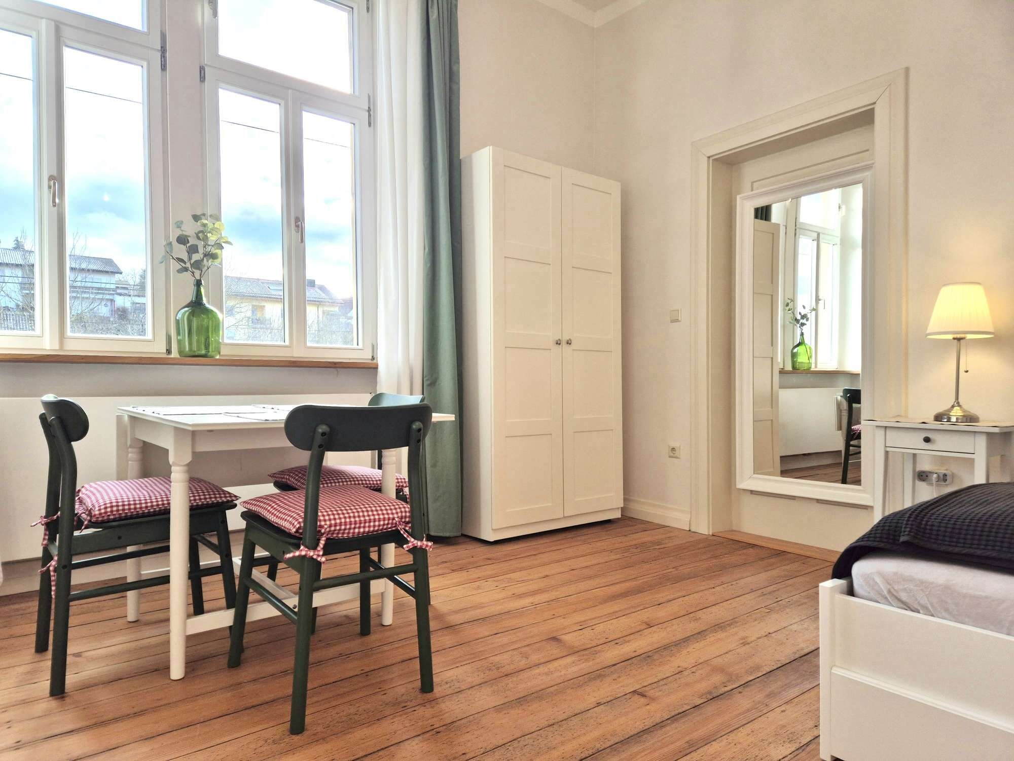 A room with a wooden floor, a white desk with two chairs, a wardrobe, a bed, and a window with curtains.