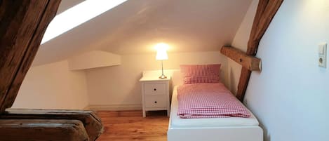 A small, cozy attic bedroom with a sloped ceiling, wooden beams, a single bed with a checkered blanket, a nightstand with a lamp, and a wooden bench.