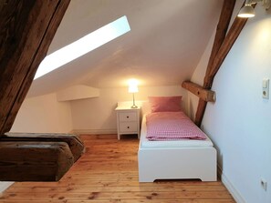 A small, cozy attic bedroom with a sloped ceiling, wooden beams, a single bed with a checkered blanket, a nightstand with a lamp, and a wooden bench.
