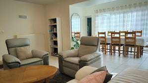 Una sala de estar con dos sillones, un sofá, una mesa de centro de madera, una estantería y un comedor con sillas y mesa de madera.