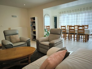 A living room with two armchairs, a sofa, a wooden coffee table, a bookshelf, and a dining area with wooden chairs and a table. - Spacious house 300 m from the sea. Sheltered garden (Luc-sur-Mer)