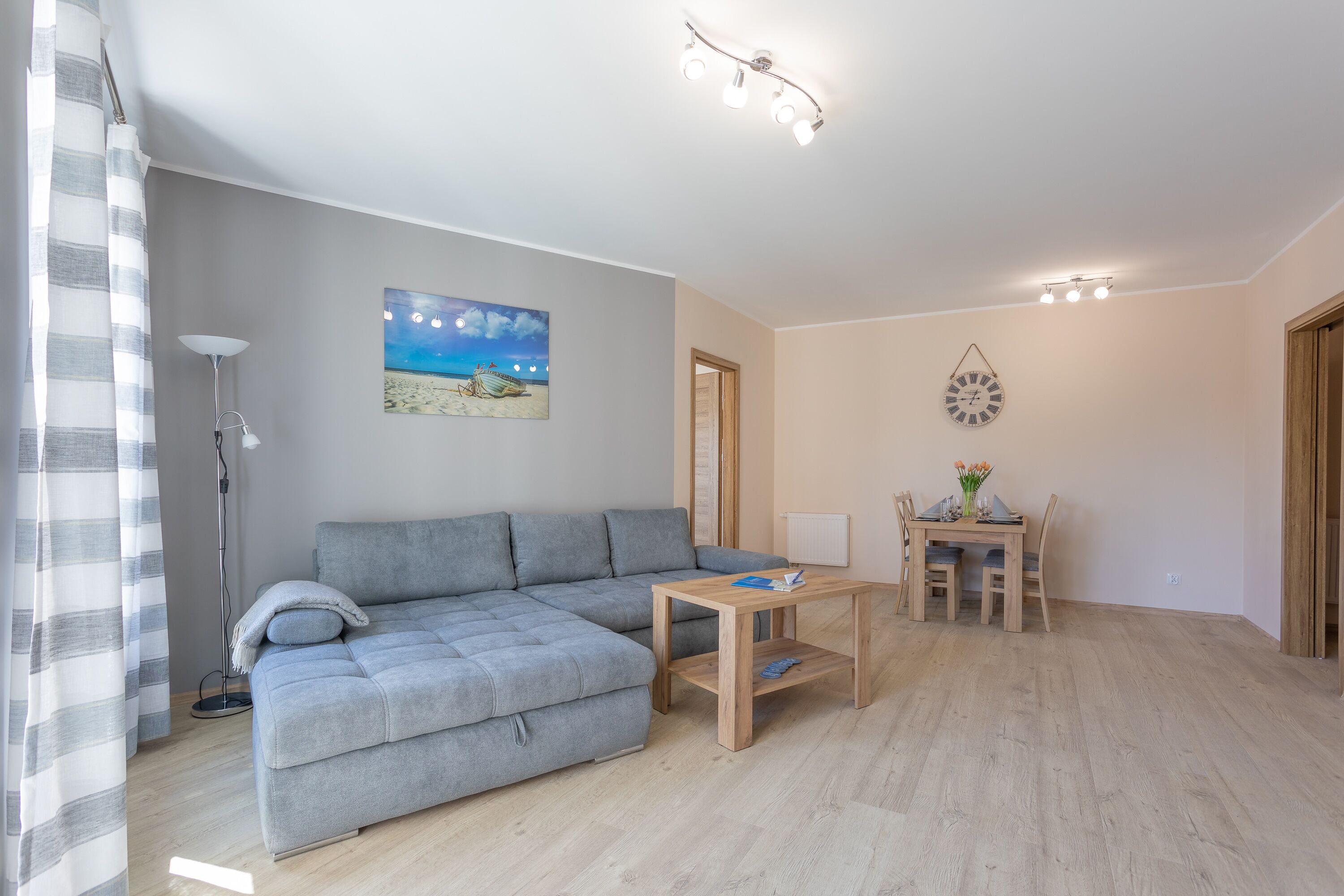 Ein modernes Wohnzimmer mit einer grauen Couch, einem Holztisch und einem Esstisch mit Stühlen. Eine Stehlampe, ein Gemälde mit Strandmotiv und eine Wanduhr sind ebenfalls vorhanden.