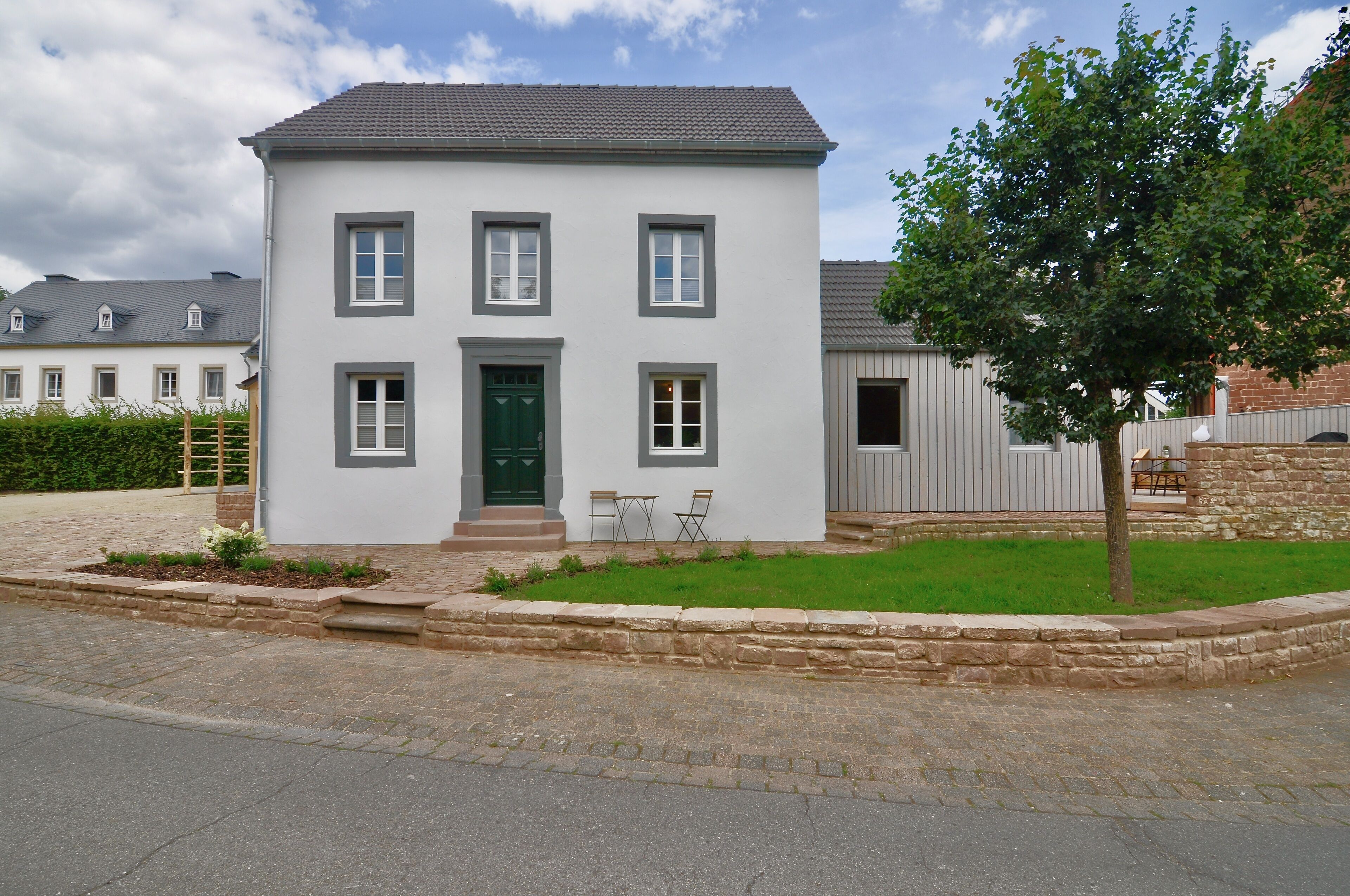 Ein zweistöckiges Haus mit grüner Tür, einer kleinen Veranda mit zwei Stühlen und einem Baum im Vorgarten.