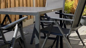 Um pátio externo com mesa e cadeiras de metal, cerca de madeira e uma pequena árvore.