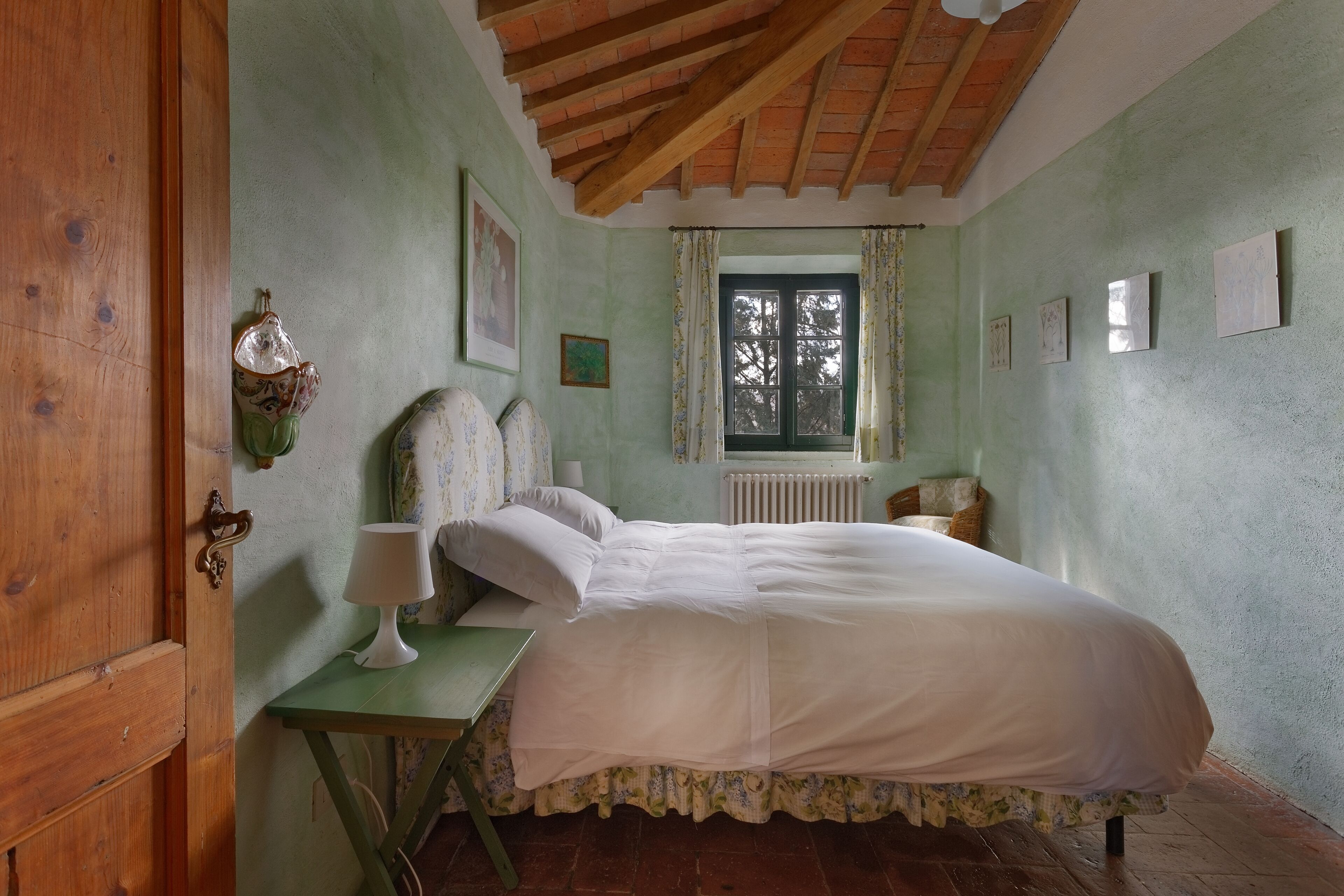 A bedroom with a wooden ceiling, a bed with white bedding, a green table with a lamp, and a window with curtains.