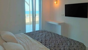 Ein Schlafzimmer mit einem Bett, einem an der Wand montierten Fernseher und einer Schiebetür, die auf einen Balkon mit Meerblick führt.